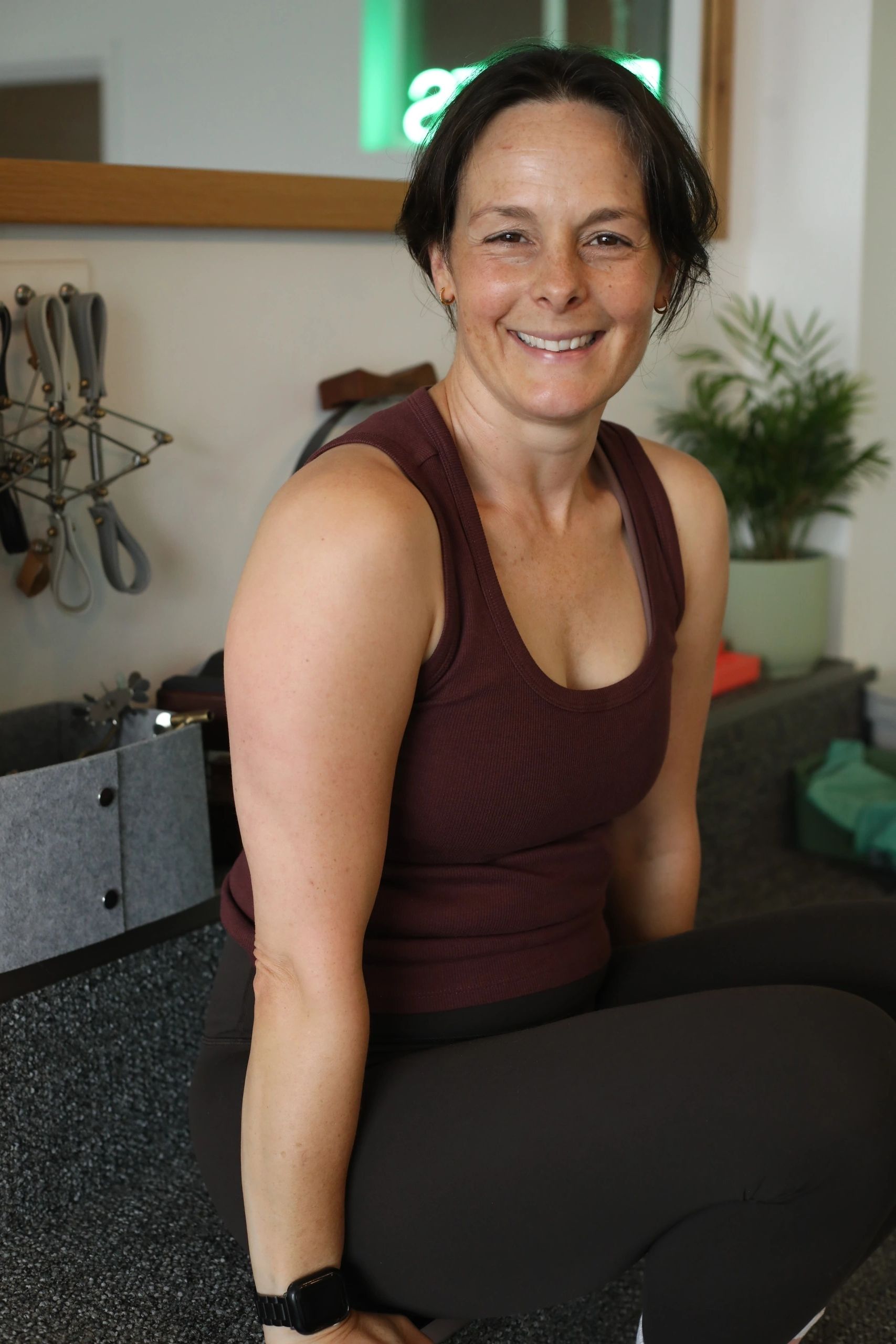 A smiling woman in a maroon tank top sitting indoors.